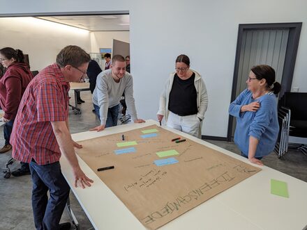 Workshop 'Digitalisierung', vier Menschen stehen diskutierend um einen Tisch mit einer als "Ideenschmiede" beschrifteten Bogen.