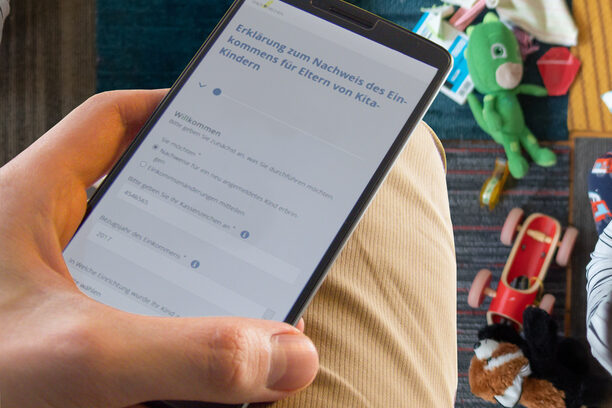 Hand hält Smartphone mit Kita-Formular, im Hintergrund spielende Kinder