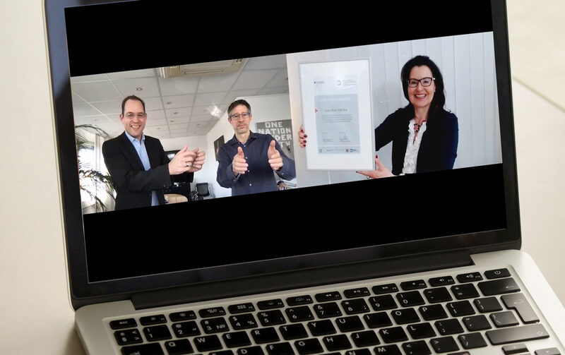 Foto eines Notebooks, auf dem Bildschirm 3 Personen in einer Videokonferenz-Situation, ein Frau hält die Urkune, 2 Männer nehmen sie lächelnd virtuell entgegen
